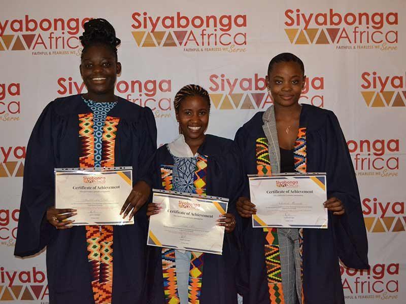Three Siyabonga Africa graduates in academic gowns holding certificates of achievement